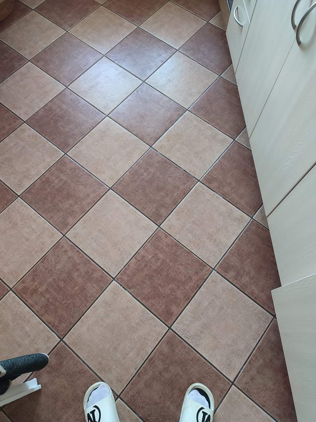 Brązowo-beżowa podłoga z płytek ułożonych w karo, widoczne białe klapki i fragment narzędzia do fugowania przy krawędzi, szafka kuchenna w tle.