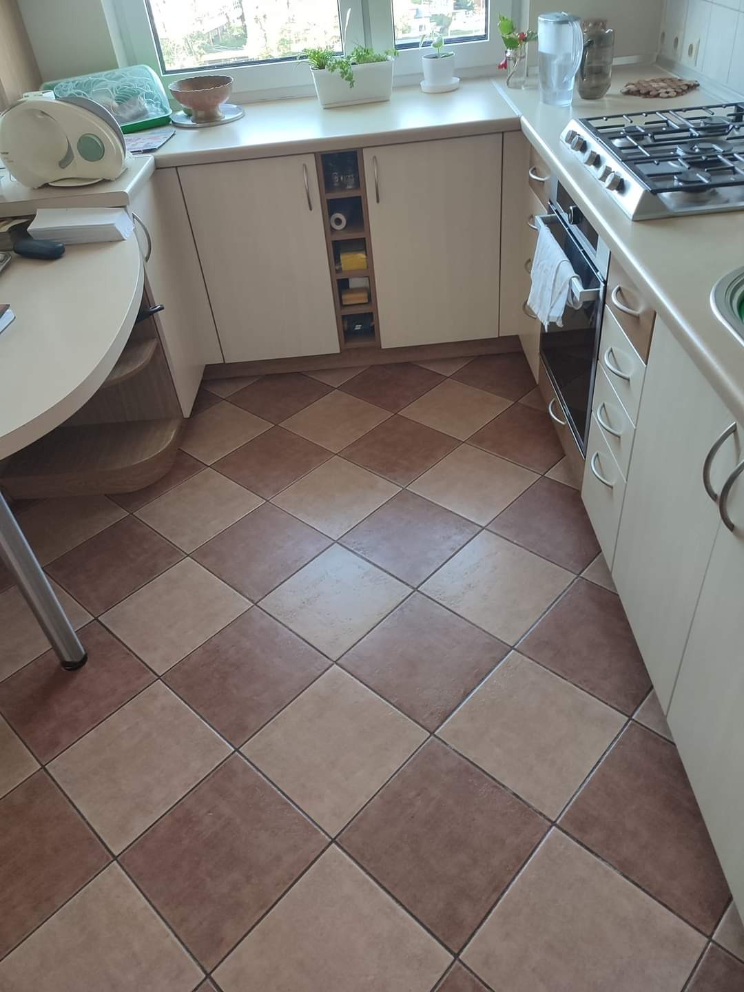 Kuchnia z brązowo-beżową podłogą w szachownicę, jasnymi szafkami i blatem, widok na okno z roślinami.