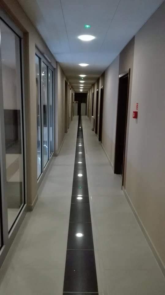 Długi, czysty korytarz z jasnymi płytkami podłogowymi i ciemnym pasem płytek z wbudowanymi punktowymi lampami LED, oświetlony sufit z okrągłymi lampami, drzwi po prawej stronie i przeszklone ściany...