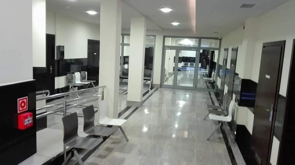 Wnętrze poczekalni z rzędem metalowych krzeseł, marmurową podłogą i białymi kolumnami, widok na szklane drzwi wejściowe.