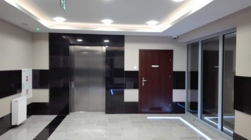 Wnętrze klatki schodowej w nowoczesnym budynku z windą, drzwiami i oknami, oświetlone ledowymi paskami podłogowymi i sufitowymi lampami.