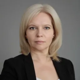 Portret kobiety o blond włosach, ubrana w czarną marynarkę, na szarym tle. Ujęcie centralne, skupione na twarzy. Widoczne delikatne cienie i naturalny odcień skóry.