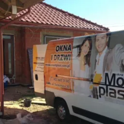 Montaż drzwi zewnętrznych w surowym budynku, widoczny samochód firmy z reklamą okien i drzwi. Pracownik w trakcie instalacji, ceglana elewacja w tle.