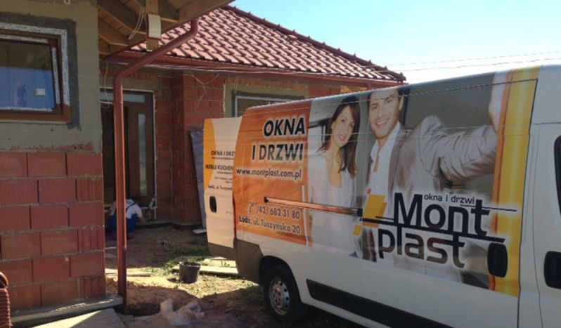 Montaż drzwi zewnętrznych w surowym budynku, widoczny samochód firmy z reklamą okien i drzwi. Pracownik w trakcie instalacji, ceglana elewacja w tle.