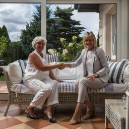 Partnerzy Agencja Nieruchomości sp. z o.o. - Dwie kobiety siedzą na wiklinowej sofie w oranżerii, uśmiechają się i ściskają dłonie. Za nimi widok na ogród z tujami i kwitnącymi hortensjami.