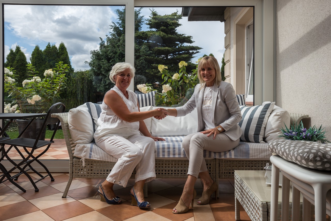 Dwie kobiety siedzą na wiklinowej sofie w oranżerii, uśmiechają się i ściskają dłonie. Za nimi widok na ogród z tujami i kwitnącymi hortensjami.