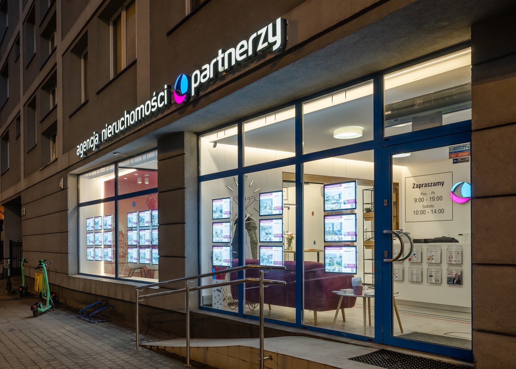 Fasada agencji nieruchomości 'Partnerzy' nocą: duże okna z wyświetlonymi ofertami, fioletowa kanapa w poczekalni, logo firmy.