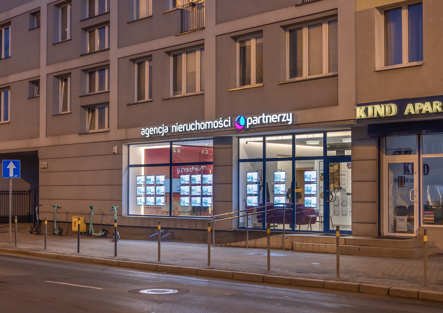 Fasada agencji nieruchomości 'partnerzy' nocą, z podświetlonym logo i witrynami pełnymi ofert sprzedaży, w tle budynek mieszkalny i zaparkowane hulajnogi.