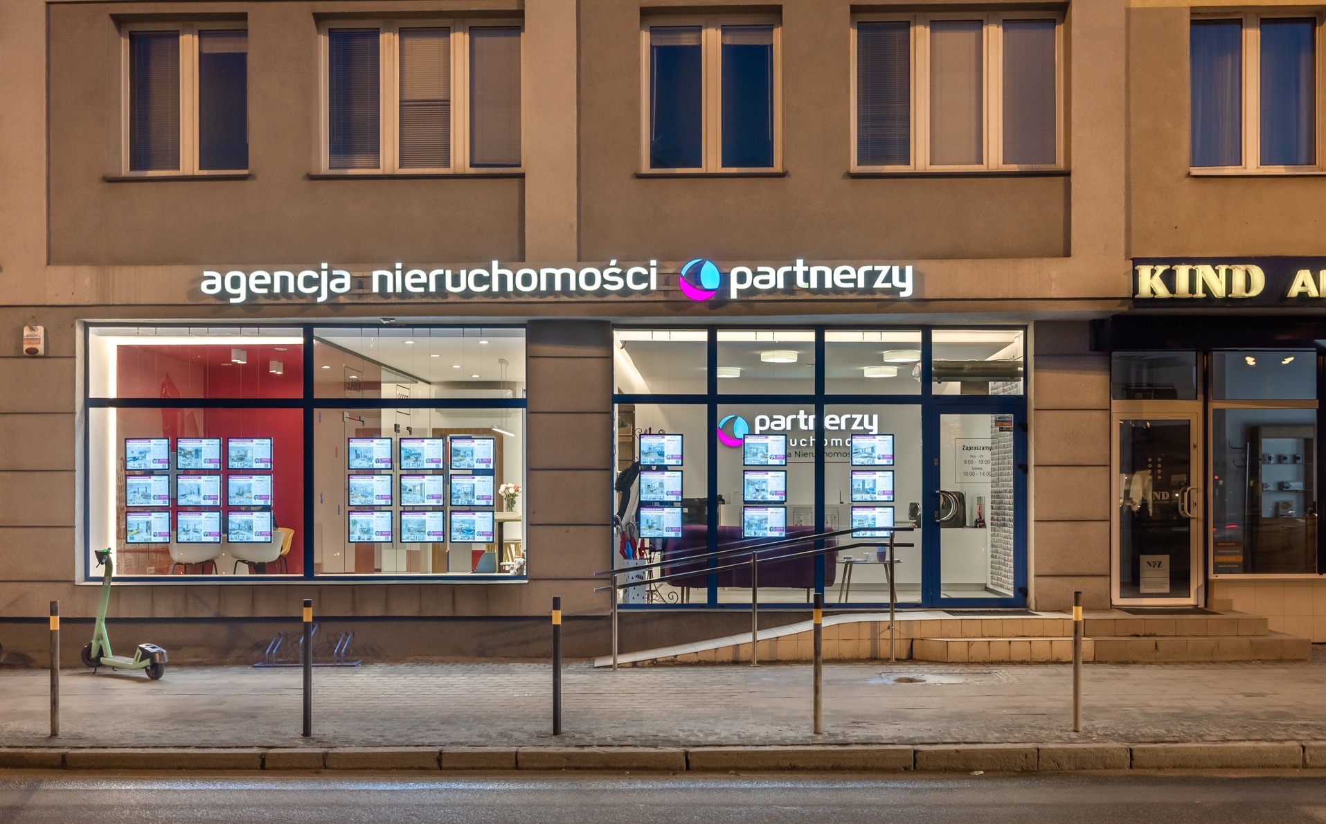 Fasada agencji nieruchomości 'partnerzy' z oświetlonym logo, wystawą ofert na monitorach i skuterem elektrycznym na chodniku. Widok nocny, miejski.