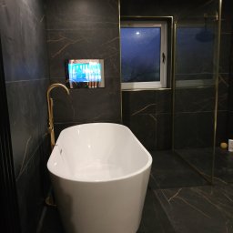 MJ serwis budowlany - Nowoczesna łazienka z wanną wolnostojącą, czarnymi płytkami z żyłkami, złotą armaturą i telewizorem wbudowanym w ścianę. Minimalistyczny design, eleganckie wykończenie.