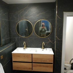 MJ serwis budowlany - Wykończona łazienka: ciemne płytki imitujące marmur, złote akcenty, podwójna umywalka z drewnianą szafką, okrągłe lustra z refleksją osoby fotografującej.