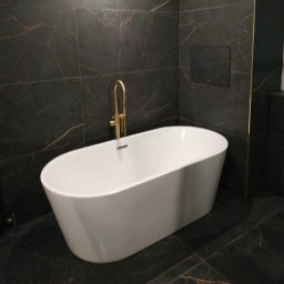 MJ serwis budowlany - Nowoczesna łazienka z wolnostojącą białą wanną i złotą armaturą na tle ciemnych płytek imitujących marmur z wyraźnym złotym żyłowaniem.