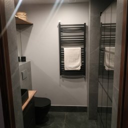 MJ serwis budowlany - Nowoczesna łazienka w szarościach z czarną armaturą: umywalka nablatowa, toaleta i grzejnik drabinkowy z ręcznikiem. Minimalistyczny design z oświetleniem punktowym.