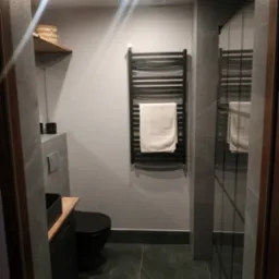 Nowoczesna łazienka w szarościach z czarną armaturą: umywalka nablatowa, toaleta i grzejnik drabinkowy z ręcznikiem. Minimalistyczny design z oświetleniem punktowym.