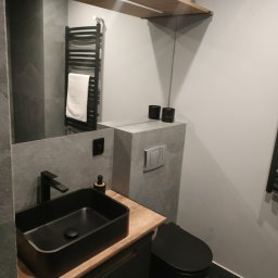 MJ serwis budowlany - Elegancka, nowoczesna łazienka w szarościach z czarną armaturą: umywalka nablatowa, toaleta podwieszana i grzejnik drabinkowy. Drewniana półka dodaje ciepła.