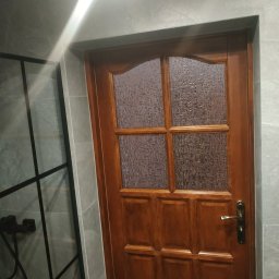 MJ serwis budowlany - Drewniane drzwi z szybą w łazience wykończonej płytkami imitującymi kamień, widoczna kabina prysznicowa z czarną armaturą i ramą. Oświetlenie górne.