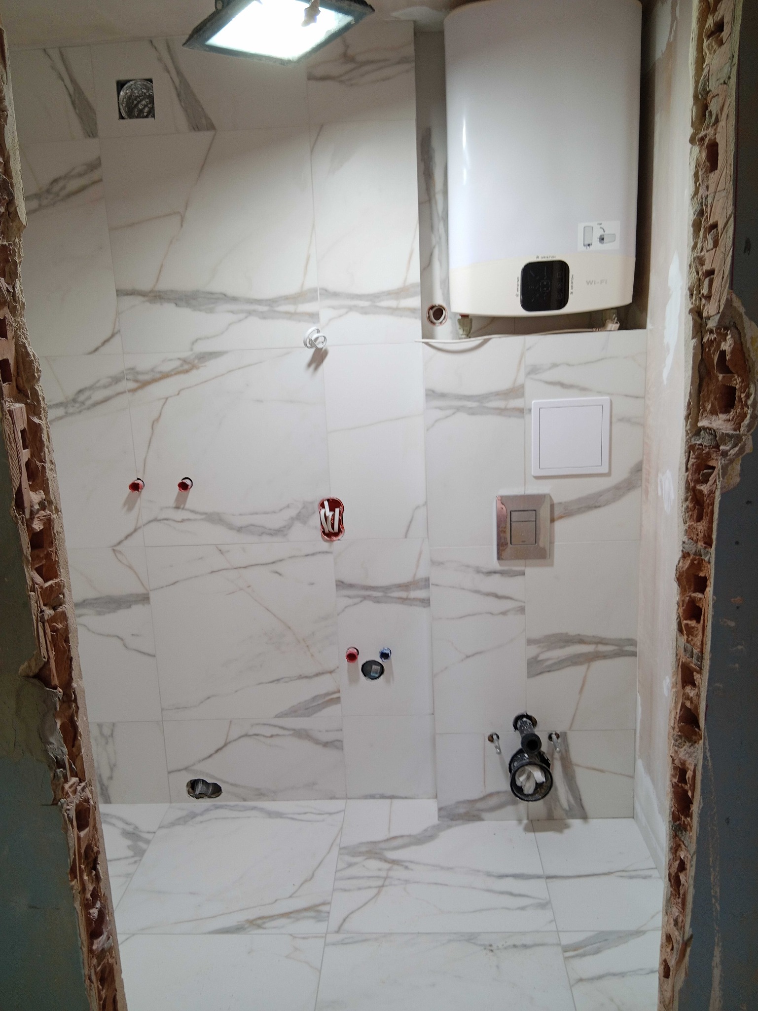 Wnętrze łazienki w trakcie remontu w Sochaczewie: białe płytki imitujące marmur na ścianach i podłodze, widoczne instalacje wodne i grzewcze, bojler na ścianie, ściany w trakcie wykończenia.