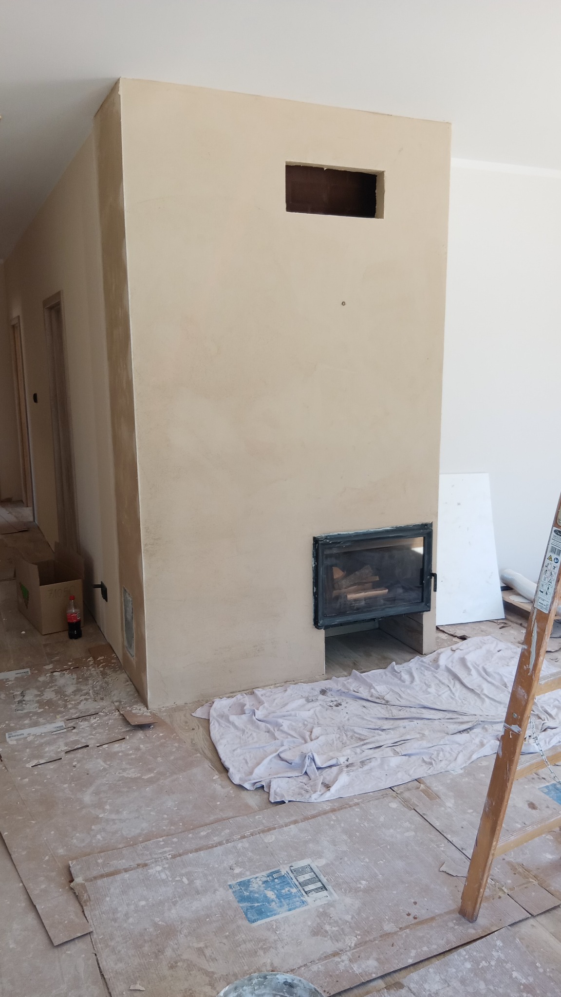 Ściana z wnęką na kominek w trakcie tynkowania, widoczne narzędzia i zabezpieczenia podłogi. Surowy stan wykończenia, beżowa kolorystyka.