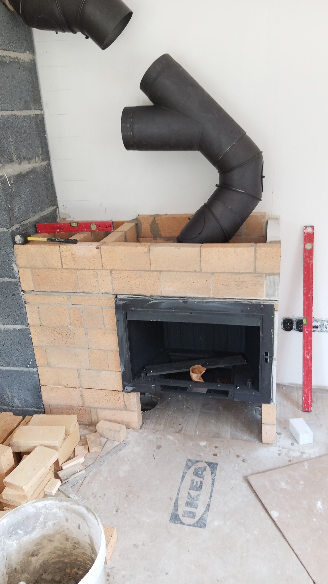 Wnętrze w trakcie budowy kominka z cegły. Widoczny wkład kominkowy, elementy przewodu dymowego i narzędzia murarskie. Na podłodze logo IKEA.