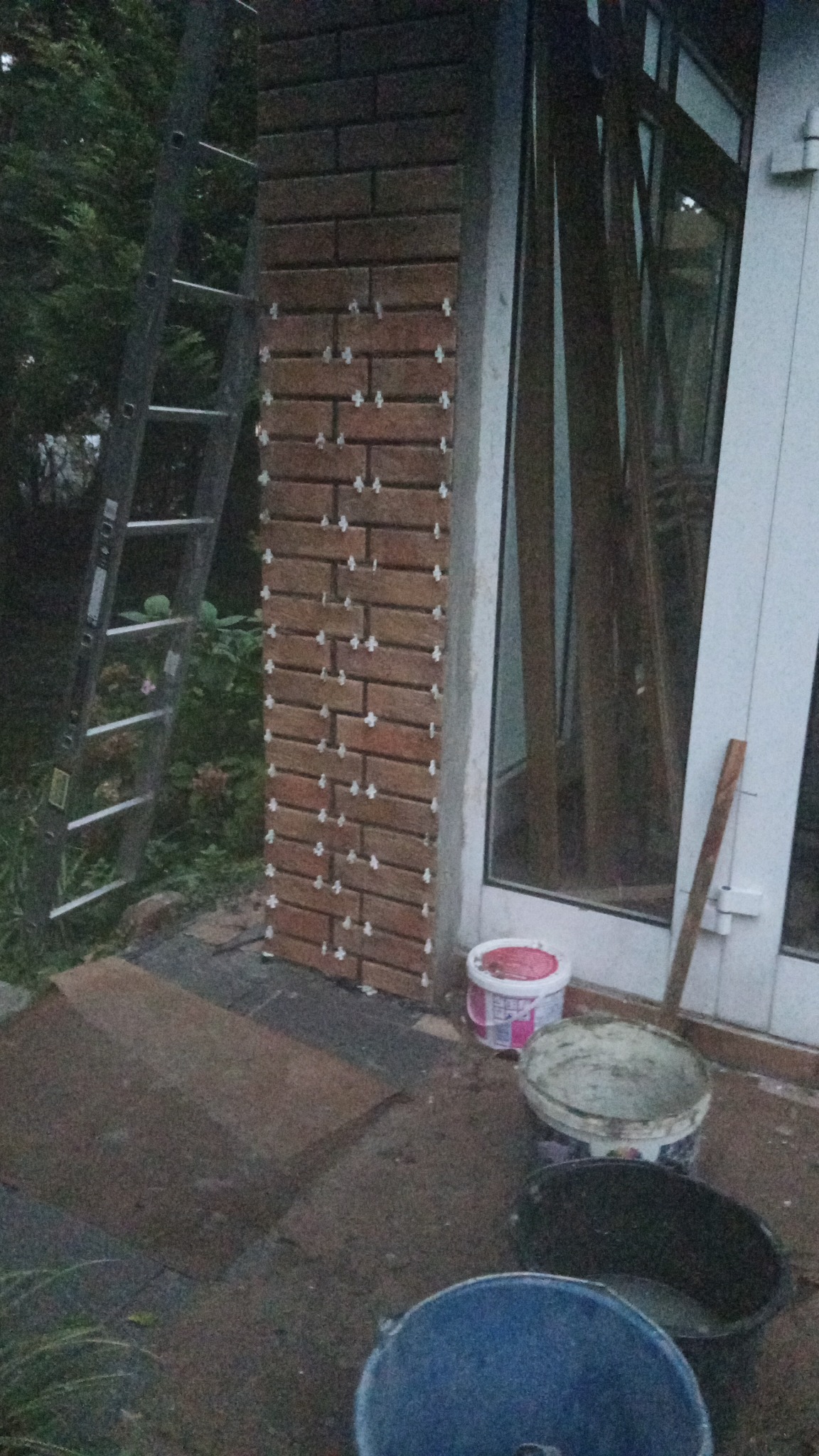 Murowanie dekoracyjnej ściany z cegieł klinkierowych przy oknie, z użyciem krzyżyków dystansowych. Widoczne wiaderka z zaprawą i drabina. Prace murarskie na zewnątrz budynku.