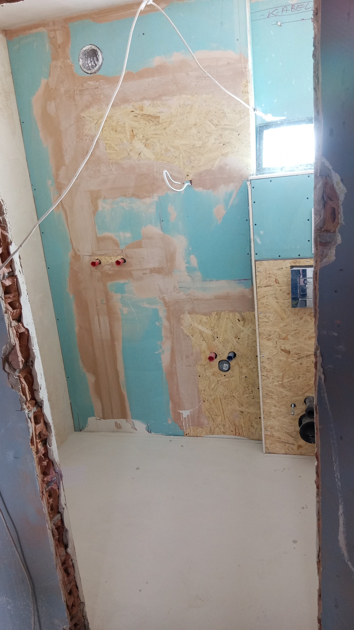 Wnętrze łazienki w trakcie remontu: płyty OSB, instalacje wodne i elektryczne, ściany z fragmentami gładzi, widoczne przewody, surowy stan wykończenia.