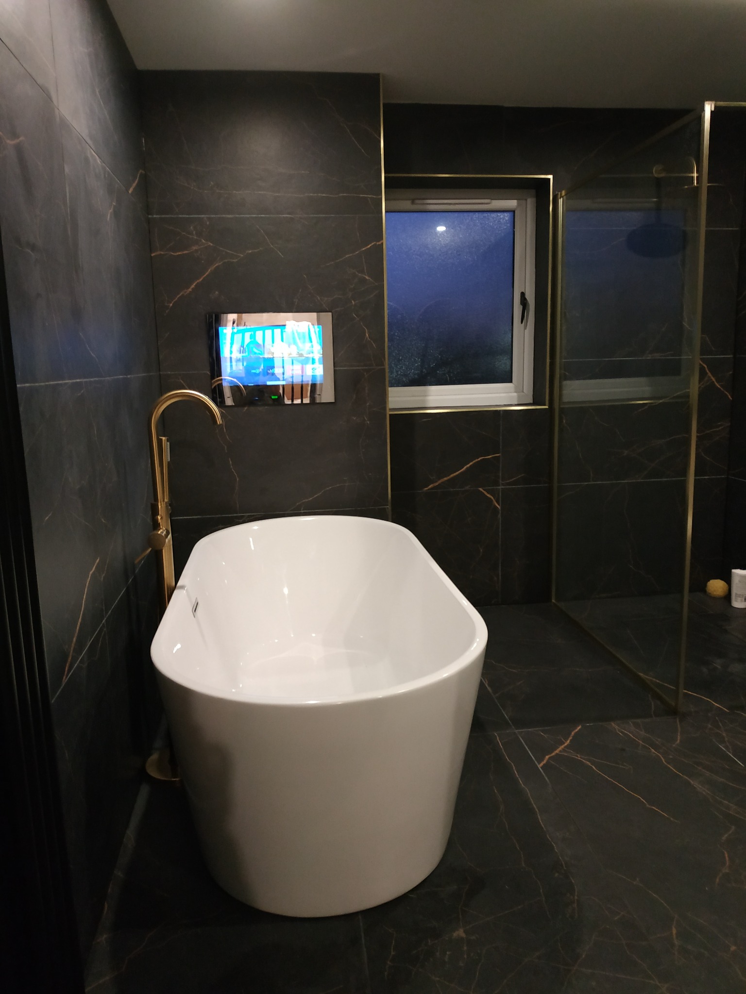 Nowoczesna łazienka z wanną wolnostojącą, czarnymi płytkami z żyłkami, złotą armaturą i telewizorem wbudowanym w ścianę. Minimalistyczny design, eleganckie wykończenie.