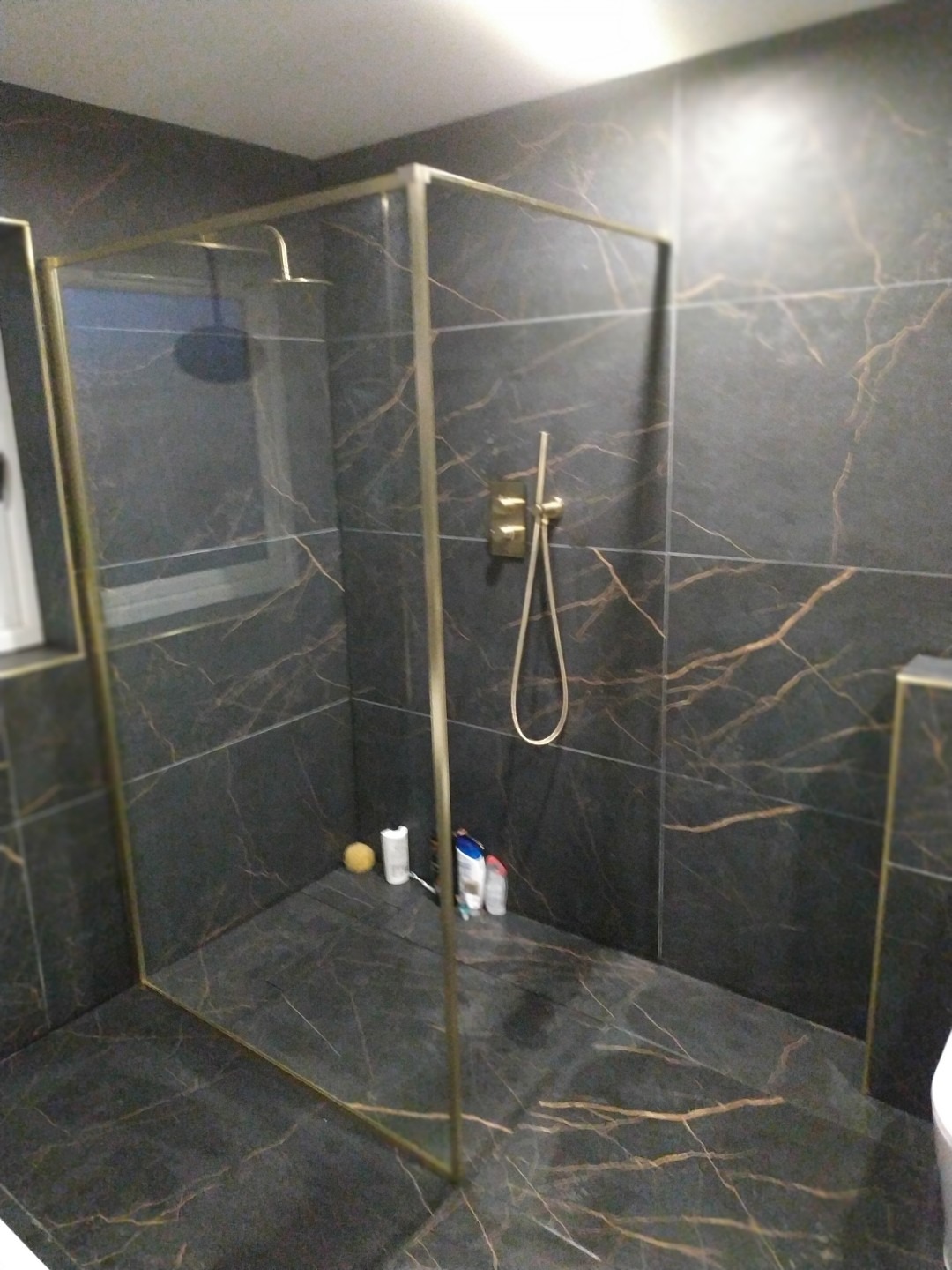 Nowoczesna kabina prysznicowa z czarnymi płytkami imitującymi marmur i złotymi profilami. Widoczne detale: bateria i akcesoria łazienkowe.