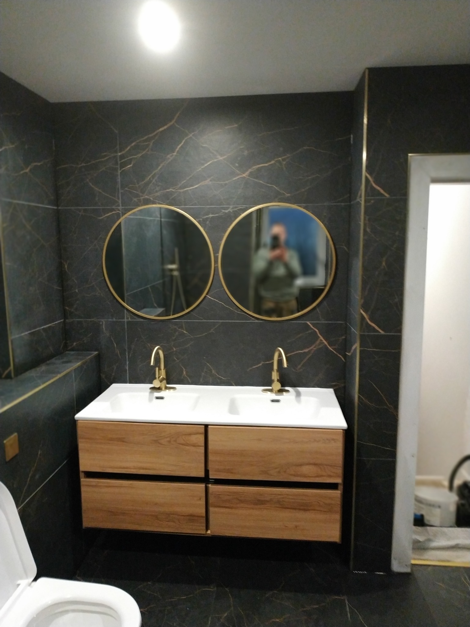 Wykończona łazienka: ciemne płytki imitujące marmur, złote akcenty, podwójna umywalka z drewnianą szafką, okrągłe lustra z refleksją osoby fotografującej.
