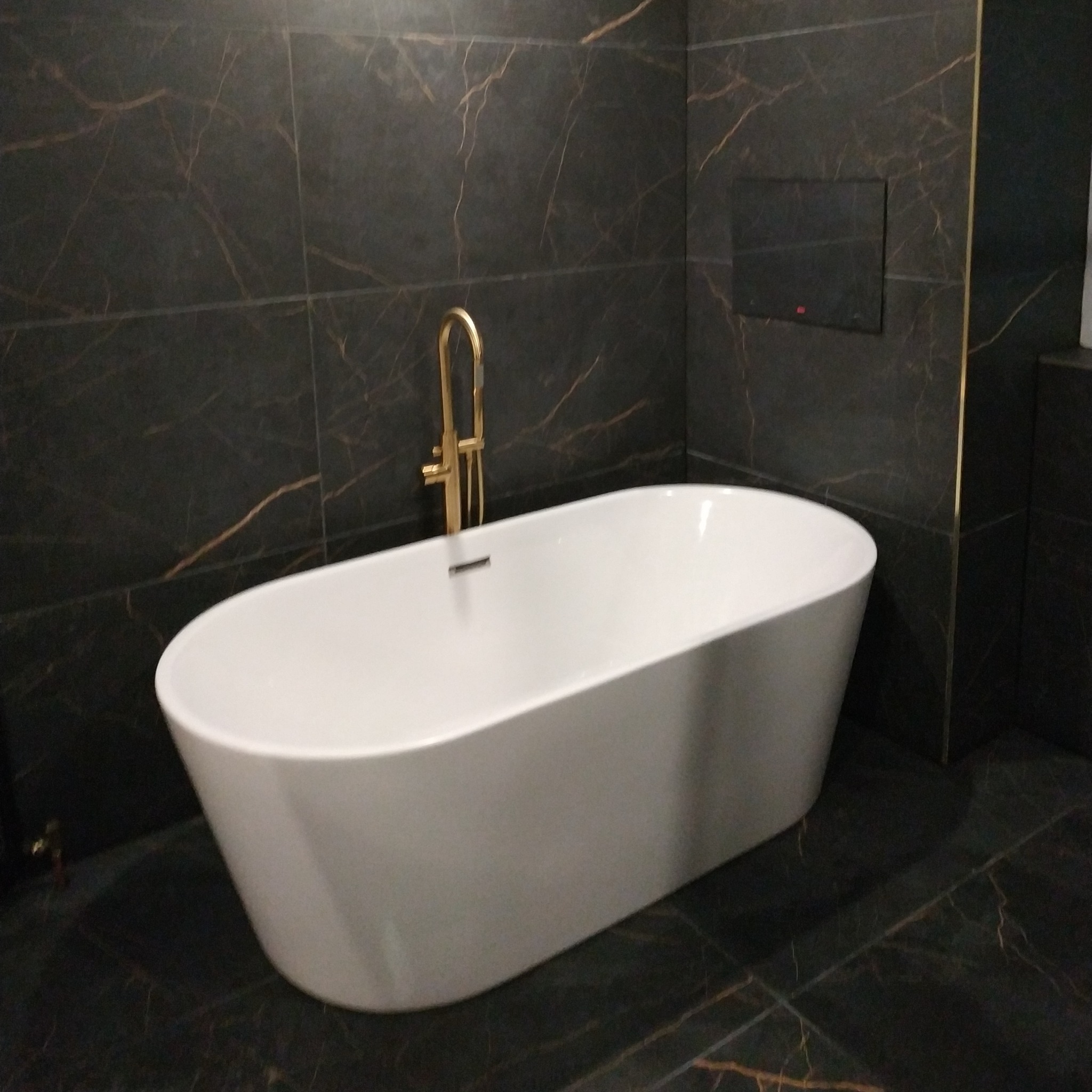 Nowoczesna łazienka z wolnostojącą białą wanną i złotą armaturą na tle ciemnych płytek imitujących marmur z wyraźnym złotym żyłowaniem.