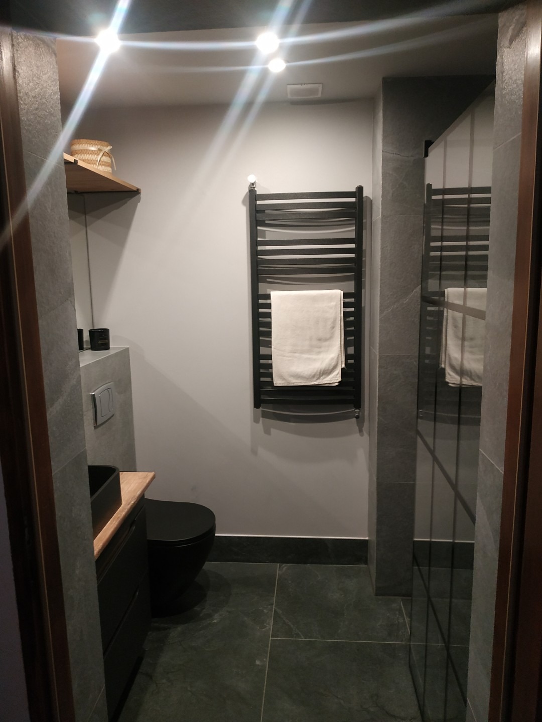 Nowoczesna łazienka w szarościach z czarną armaturą: umywalka nablatowa, toaleta i grzejnik drabinkowy z ręcznikiem. Minimalistyczny design z oświetleniem punktowym.