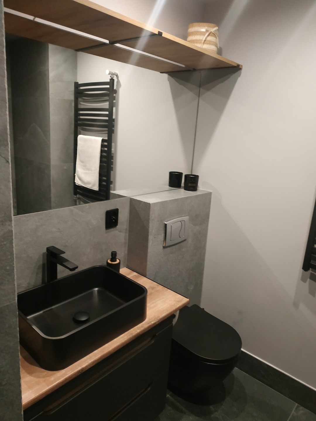 Elegancka, nowoczesna łazienka w szarościach z czarną armaturą: umywalka nablatowa, toaleta podwieszana i grzejnik drabinkowy. Drewniana półka dodaje ciepła.