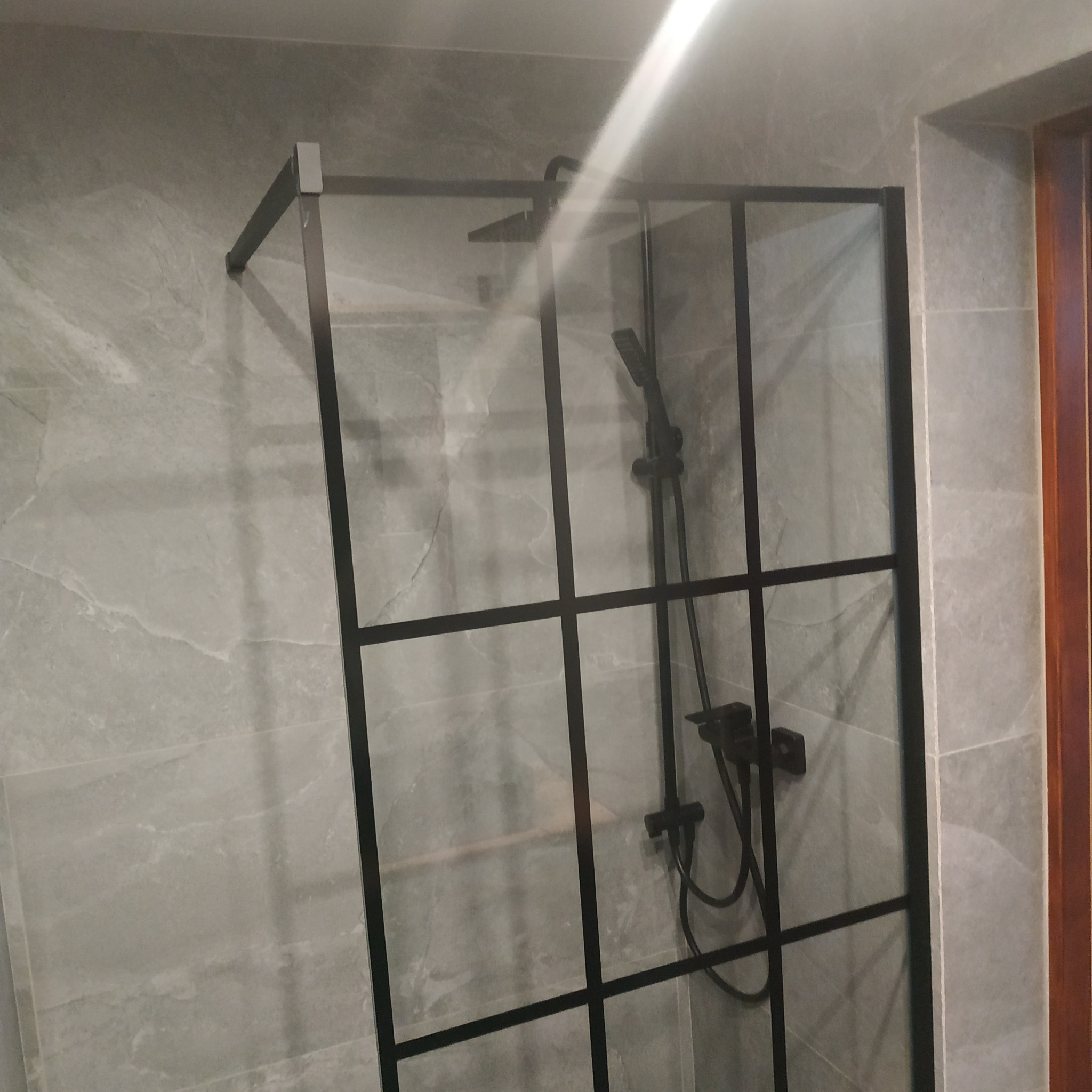 Kabina prysznicowa z czarnymi profilami na tle szarych płytek imitujących marmur. Widoczna deszczownica i bateria. Minimalistyczny design.