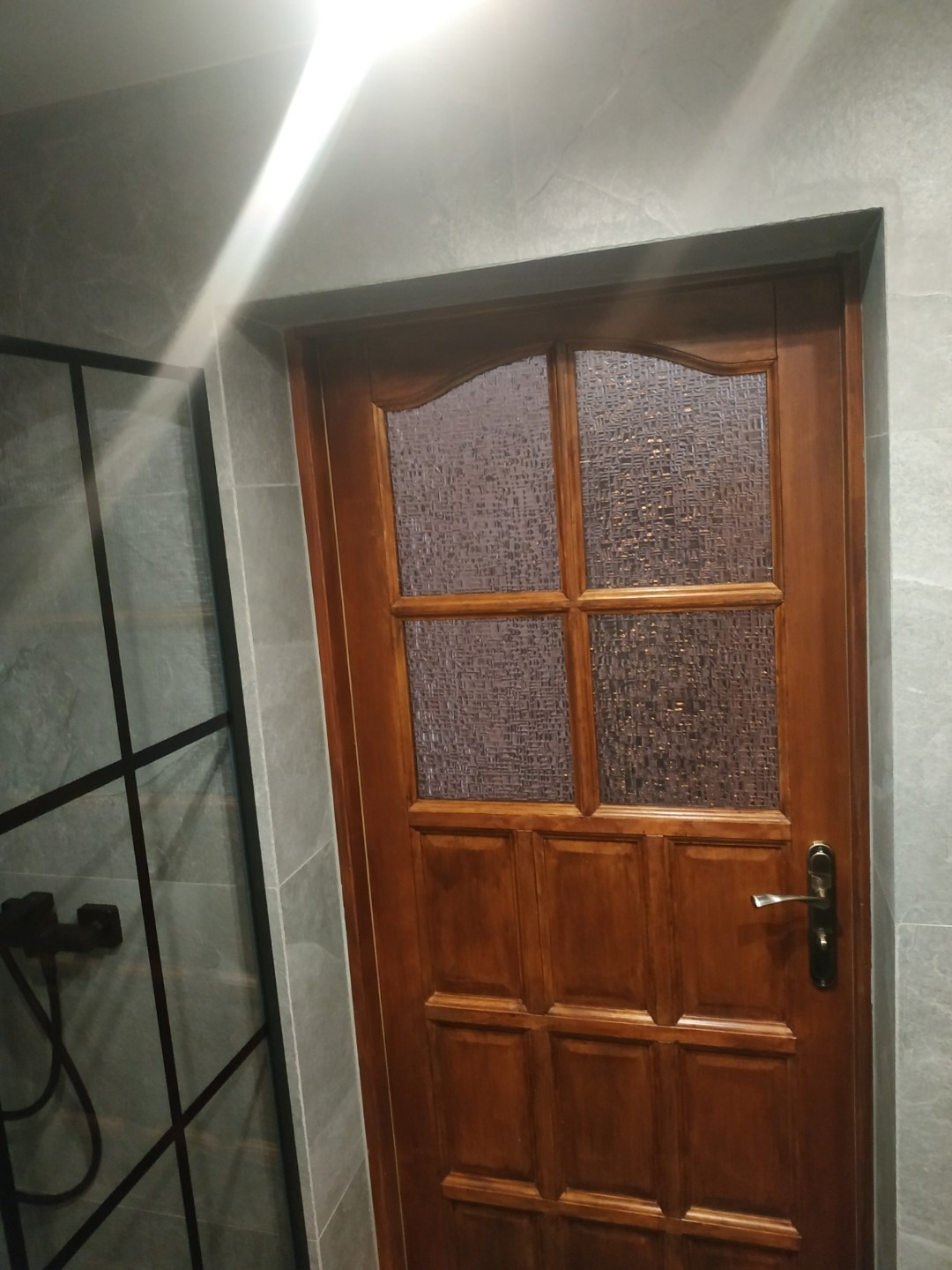 Drewniane drzwi z szybą w łazience wykończonej płytkami imitującymi kamień, widoczna kabina prysznicowa z czarną armaturą i ramą. Oświetlenie górne.