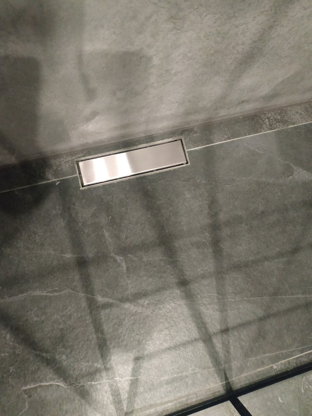 Nowoczesny odpływ liniowy w szarej, wykończonej łazience, z widocznym fragmentem kabiny prysznicowej. Minimalistyczny design i dbałość o detale.