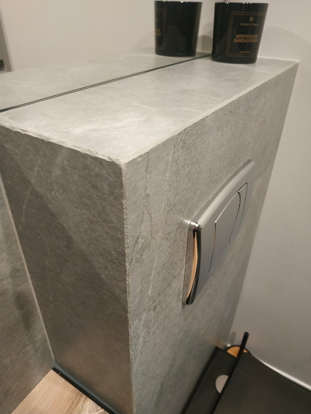 Szara zabudowa toalety podtynkowej z chromowanym przyciskiem spłukującym i dwoma świecami zapachowymi na górze.  Wykończenie imituje kamień.