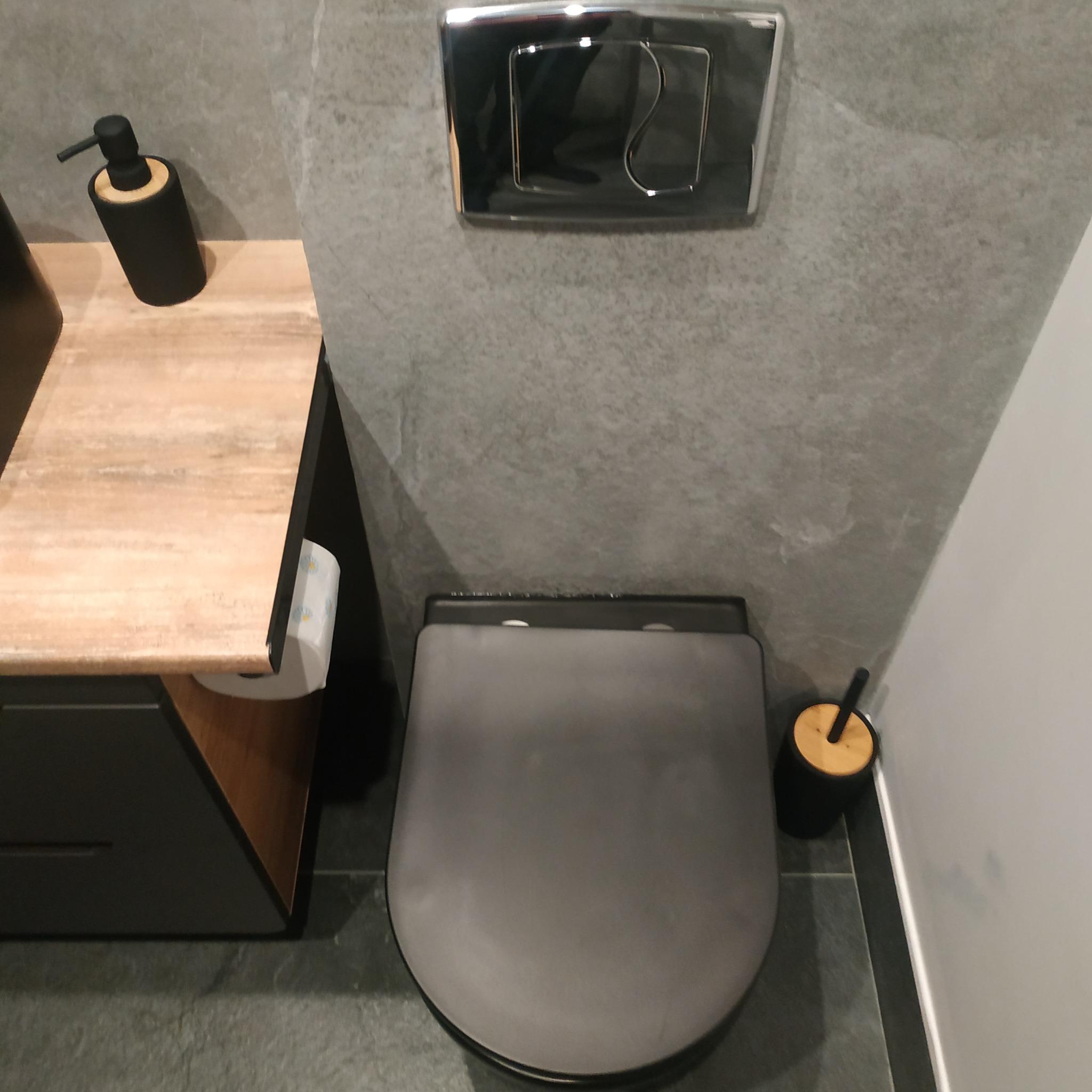 Nowoczesna łazienka w odcieniach szarości z podwieszaną toaletą, chromowanym przyciskiem spłukującym i drewnianym akcentem na blacie umywalki.