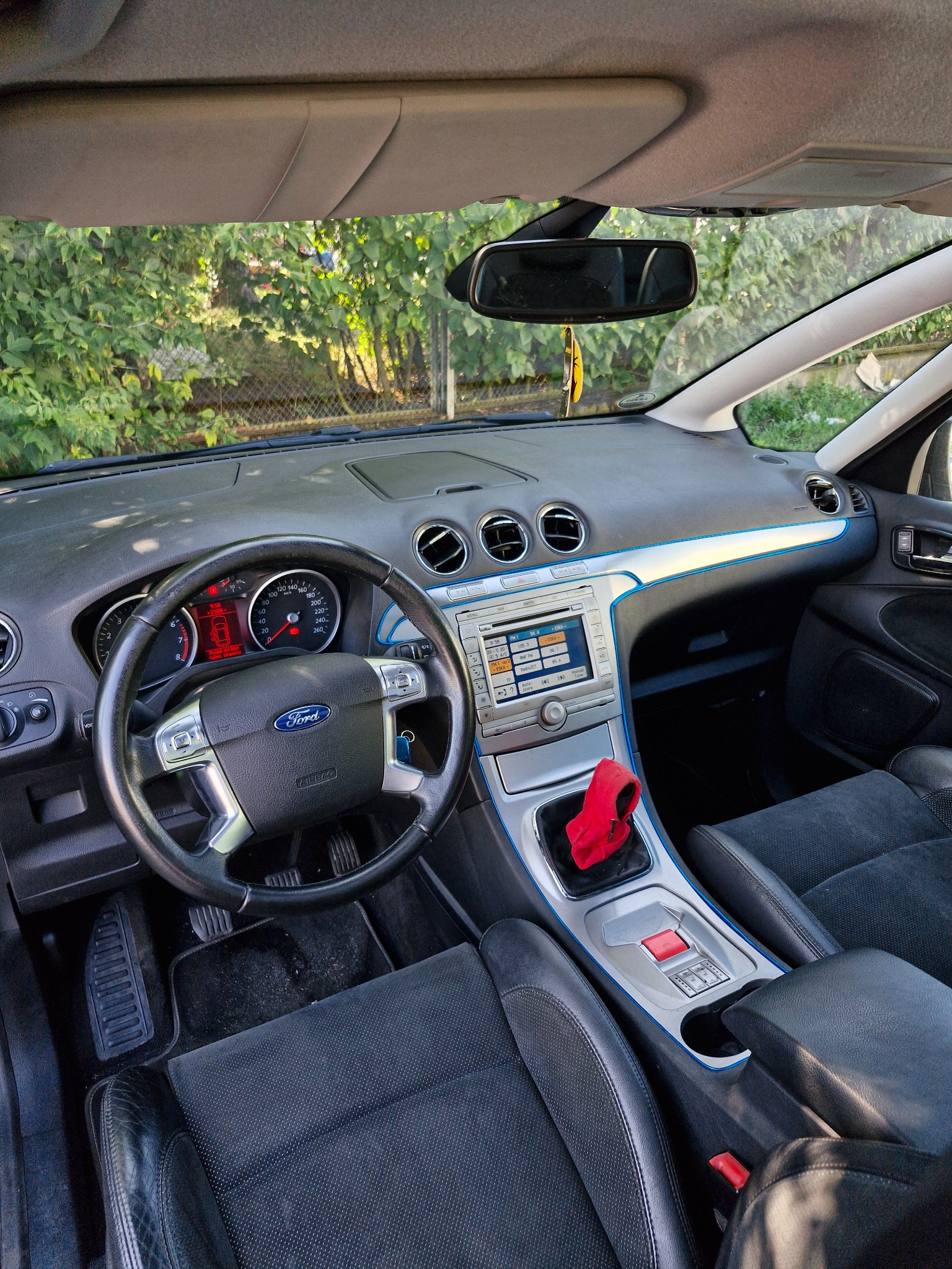 Wnętrze samochodu Ford S-Max z widoczną deską rozdzielczą, kierownicą z logo Forda, radiem samochodowym oraz czarnymi skórzanymi fotelami. Na dźwigni zmiany biegów leży czerwona ściereczka.