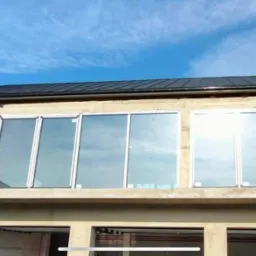 Budynek w trakcie montażu okien, widoczne rusztowanie i częściowo surowy stan elewacji, na górze rząd okien z białymi ramami, odbijających błękitne niebo.