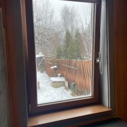 Calmex24 Cezary Okraska - Brązowe okno z roletą zwiniętą u góry, widok na zasypany śniegiem ogród z drewnianym płotem.
