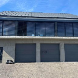 Calmex24 Cezary Okraska - Nowoczesny budynek z trzema szarymi bramami garażowymi, szarymi roletami zewnętrznymi na piętrze i drzwiami wejściowymi z boku, na żwirowym podłożu.