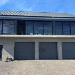 Nowoczesny budynek z trzema szarymi bramami garażowymi, szarymi roletami zewnętrznymi na piętrze i drzwiami wejściowymi z boku, na żwirowym podłożu.