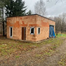 Surowy budynek z pustaków z zamontowanymi oknami i drzwiami wejściowymi, przykryty plandeką otwór garażowy, jesienne otoczenie.