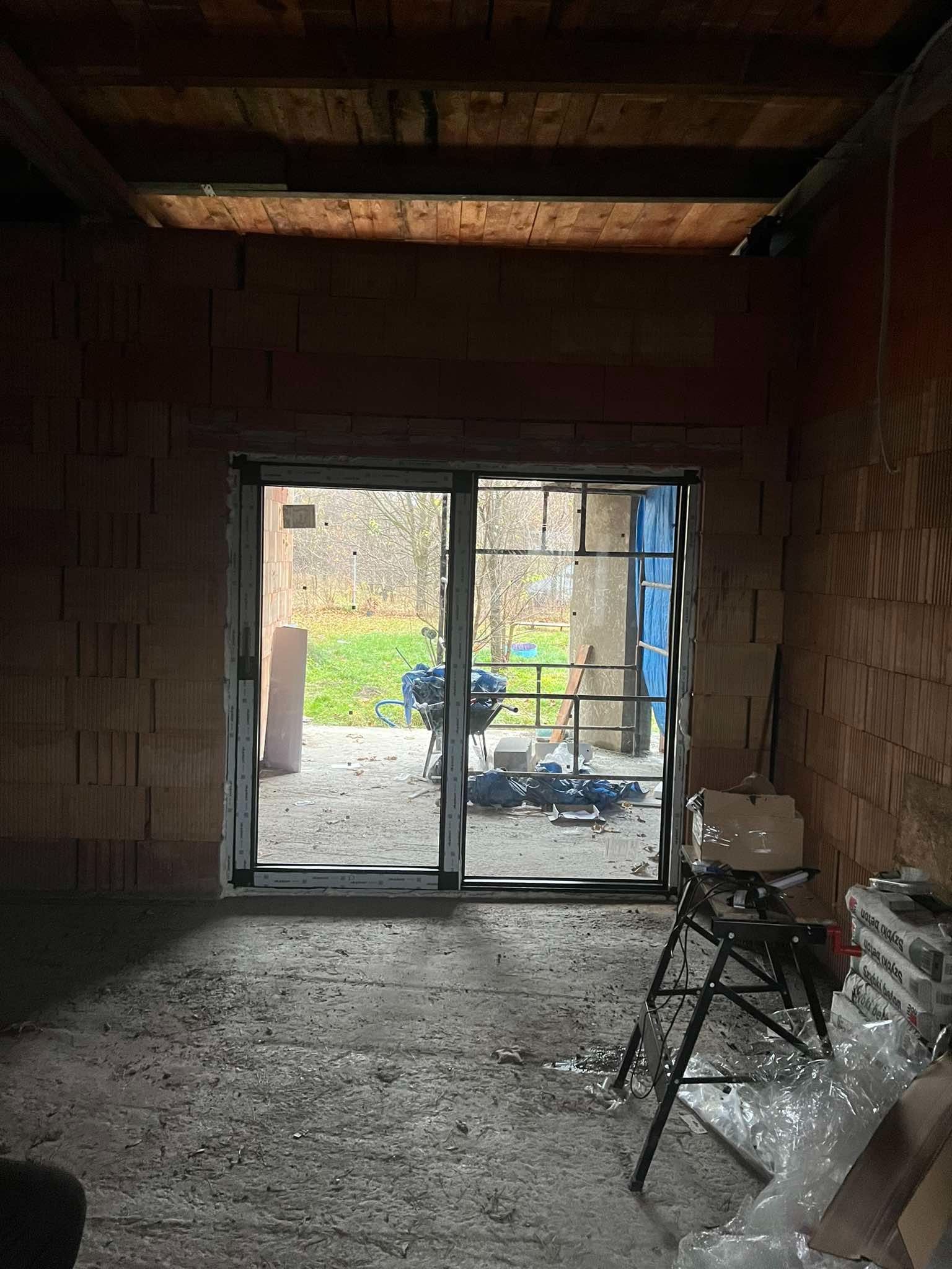 Surowe wnętrze domu w trakcie budowy z widokiem na ogród przez przesuwne okno. Na podłodze widoczne ślady po budowie, a w tle narzędzia i materiały budowlane.