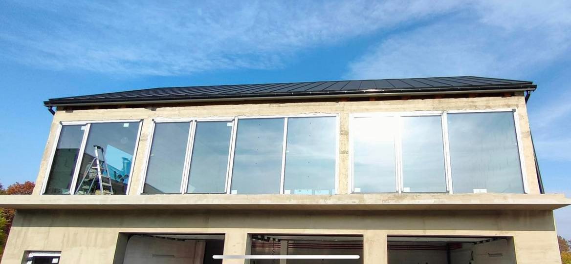 Budynek w trakcie montażu okien, widoczne rusztowanie i częściowo surowy stan elewacji, na górze rząd okien z białymi ramami, odbijających błękitne niebo.