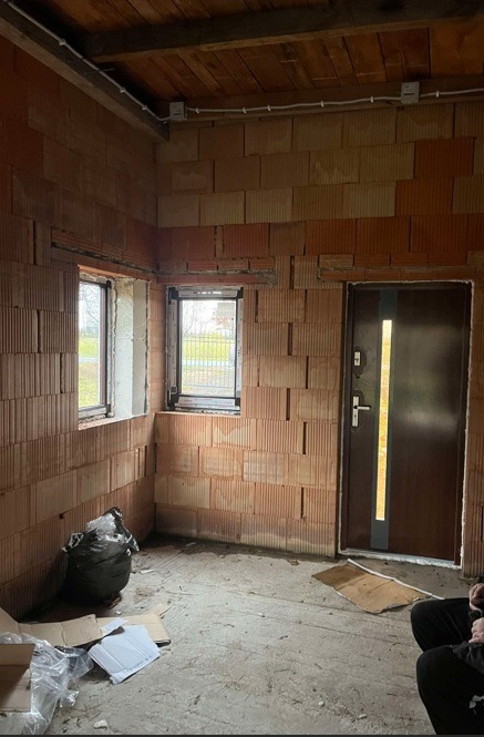 Surowy stan wnętrza z brązową stolarką okienną i drzwiami zewnętrznymi z metalową wstawką, widoczne niedokończone ściany z pustaków, instalacja elektryczna pod sufitem i prowizoryczne...