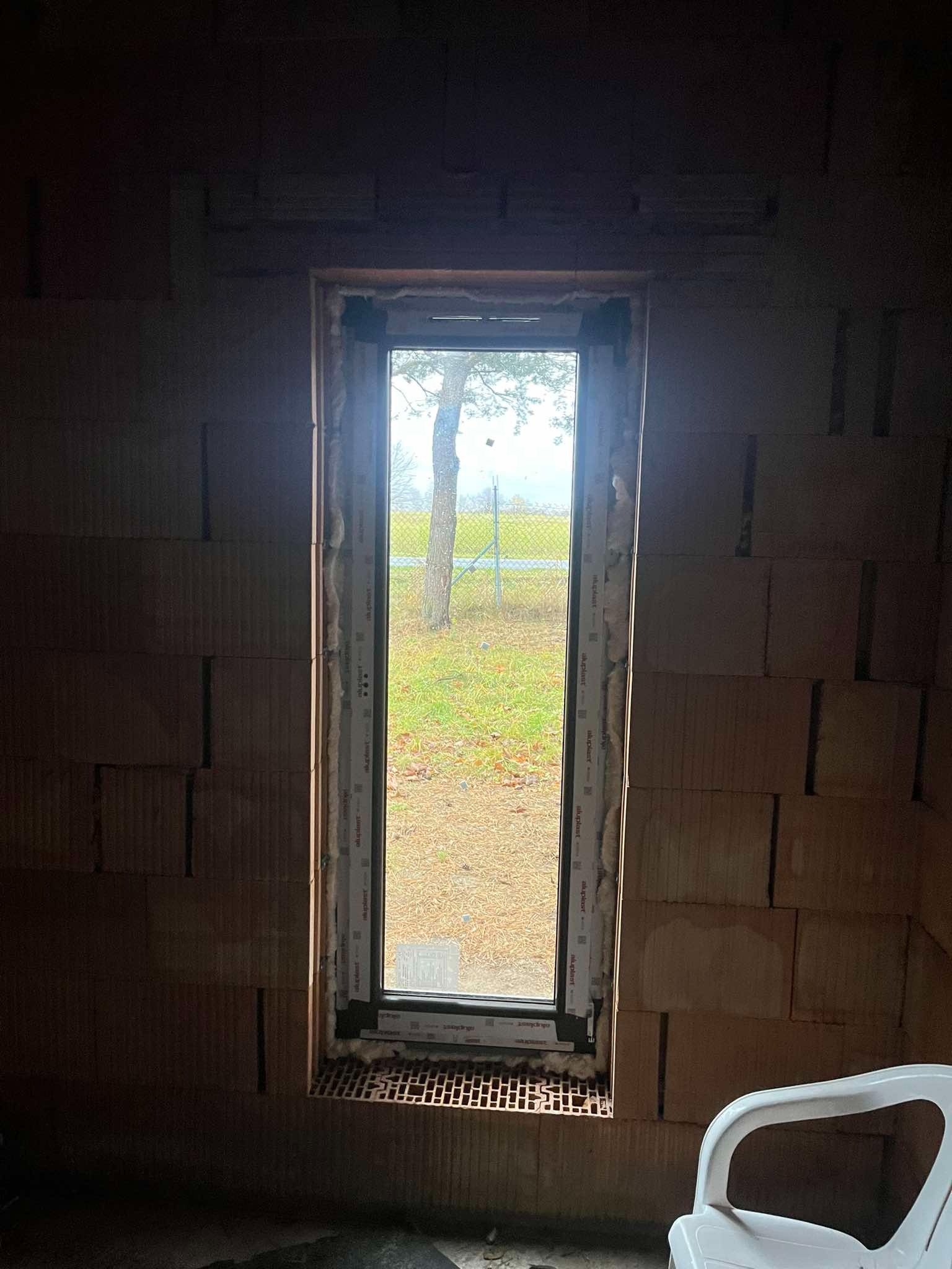 Montaż okna w surowym stanie; widok na zewnątrz przez pojedyncze okno w trakcie instalacji, z widoczną pianką montażową i częściowo wykończonym murem z cegieł.