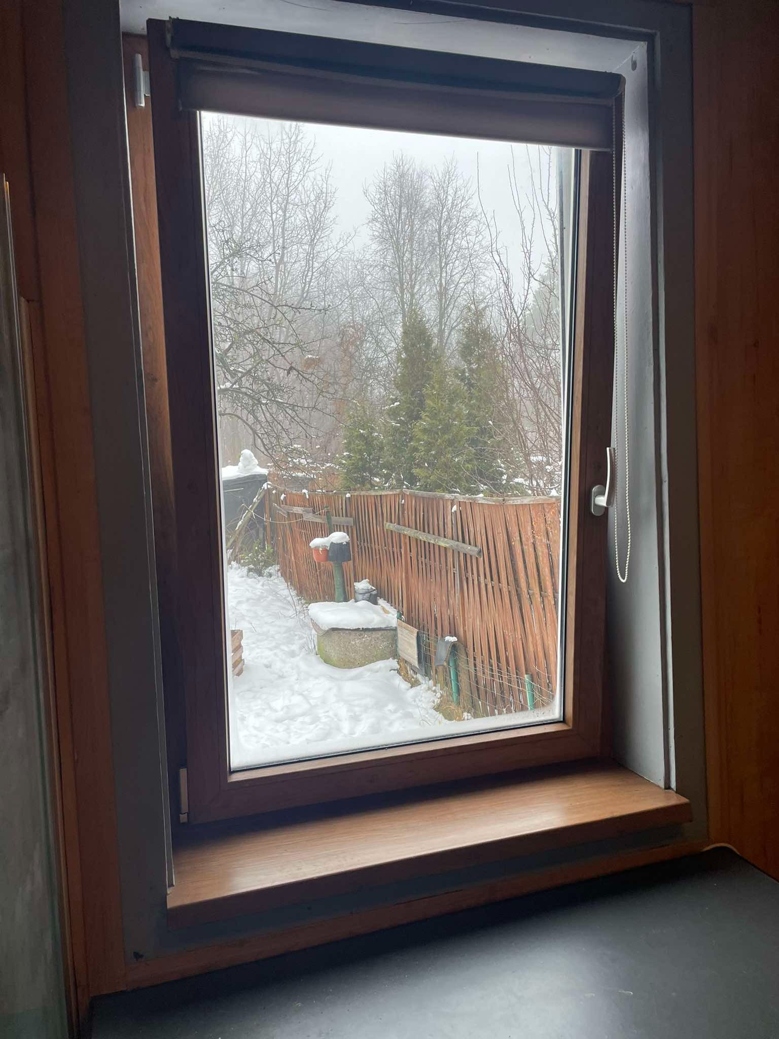 Brązowe okno z roletą zwiniętą u góry, widok na zasypany śniegiem ogród z drewnianym płotem.