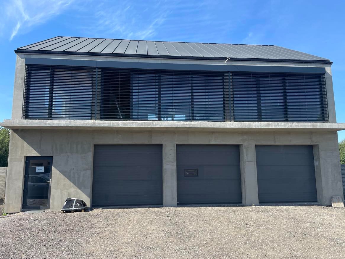 Nowoczesny budynek z trzema szarymi bramami garażowymi, szarymi roletami zewnętrznymi na piętrze i drzwiami wejściowymi z boku, na żwirowym podłożu.