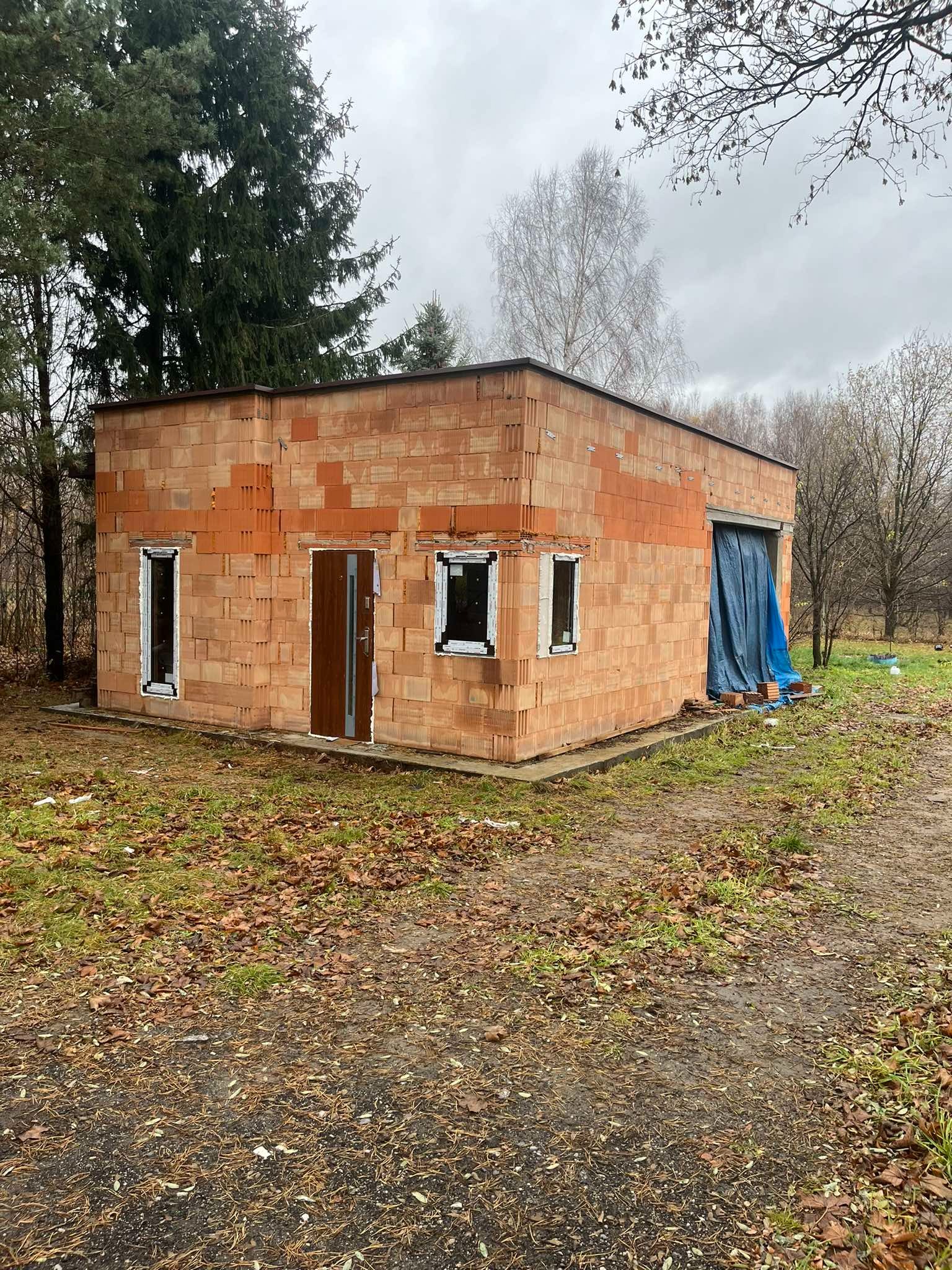 Surowy budynek z pustaków z zamontowanymi oknami i drzwiami wejściowymi, przykryty plandeką otwór garażowy, jesienne otoczenie.