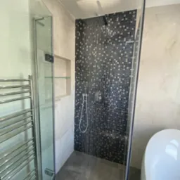 Nowoczesna kabina prysznicowa z mozaiką w odcieniach szarości i czerni, szklane drzwi, grzejnik drabinkowy i fragment białej wanny w tle.